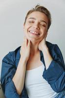 A young woman sitting in a chair at home smiling with teeth with a short haircut in jeans and a denim shirt on a white background. Girl natural poses with no filters photo