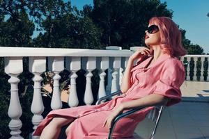 a glamorous woman in sunglasses in the summer sits on the street on a chair near the railing photo