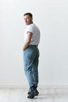 Full body view from the back shot of handsome serious tanned man guy in basic t-shirt looks at camera posing on white background. Fashion Style New Collection Offer. Copy space for ad. Model snap photo