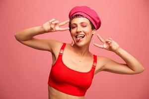 Young athletic woman with a short haircut and purple hair in a red top and a pink hat with an athletic figure smiles and grimaces looking at the camera on a pink background photo