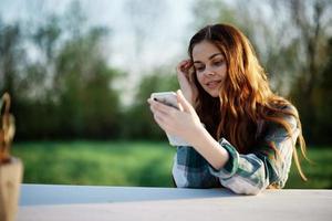 joven niña blogger persona de libre dedicación participación su teléfono mirando a eso y trabajando al aire libre en un verde parque pensativamente mirando a el pantalla en línea foto