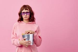 Cool movie. Confused cute redhead lady in pink hoodie sunglasses with popcorn posing isolated on over pink studio background show copy space Banner. Fashion Cinema concept. Entertainment offer photo