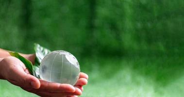 Close up of hands holding crystal globe on green grass with blur nature background, earth day or world environment day concept. Green world and sustainable conservation of forest resources. photo
