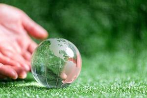 cerca arriba de manos con cristal globo en verde césped con difuminar naturaleza fondo, tierra día o mundo ambiente día concepto. verde mundo y sostenible conservación de bosque recursos. foto