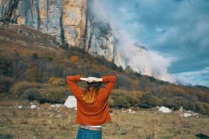 un mujer en un suéter sostiene su manos detrás su cabeza y mira a el alto montañas paisaje azul cielo nubes foto