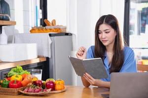 A young woman with a beautiful face in a blue shirt with long hair eating fruit sitting inside the kitchen at home with a laptop and notebook for relaxation, Concept Vacation. photo