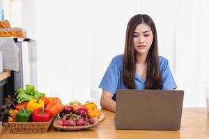 un joven mujer con un hermosa cara en un azul camisa con largo pelo comiendo Fruta sentado dentro el cocina a hogar con un ordenador portátil y cuaderno para relajación, concepto vacaciones. foto