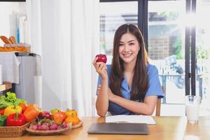 A young woman with a beautiful face in a blue shirt with long hair eating fruit sitting inside the kitchen at home with a laptop and notebook for relaxation, Concept Vacation. photo