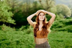 un joven mujer con un atlético cuerpo camina en un verano verde parque en naturaleza. puesta de sol luz de sol ilumina su rojo pelo foto