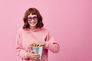 Enjoyed happy smiling cute redhead lady in pink hoodie sunglasses with popcorn posing isolated on over pink studio background. Copy space Banner. Fashion Cinema Movie concept. Entertainment offer photo