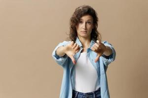 Unhappy curly beautiful female in jeans casual shirt show thumbs down gesture Dislike at camera posing isolated on over beige pastel background. People Lifestyle emotions concept. Copy space photo