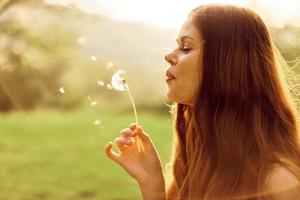 Portrait of a young woman in profile with a dandelion flower in her hand blowing on it and smiling against the green summer grass in the setting sun in nature photo