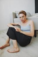 Teenage girl sitting on the floor at home smiling in home clothes and glasses with a short haircut, lifestyle without filters, free copy space photo