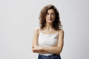 Pensive tricky smiling curly beautiful woman in basic white t-shirt looks aside posing isolated on over white background. People Emotions Lifestyle concept. Copy space. Closeup portrait in studio photo