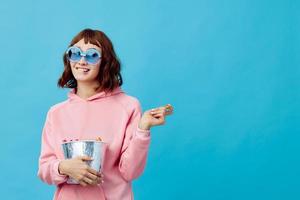 Evening movie concept. Lovely smiling cute redhead lady in pink hoodie sunglasses with popcorn posing isolated on blue studio background. Copy space Banner. Fashion Cinema. Entertainment offer photo