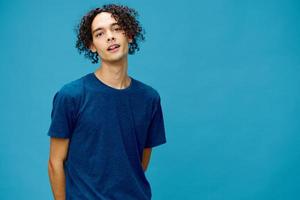 Smiling joyful cute curly tanned Caucasian guy in basic t-shirt posing isolated on over blue background. Lifestyle and Emotions concept. Good offer with copy space for ad photo