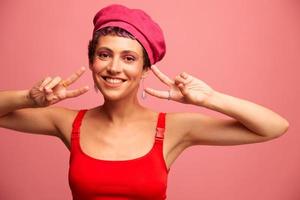 joven atlético mujer con un corto Corte de pelo y púrpura pelo en un rojo parte superior y un rosado sombrero con un atlético figura sonrisas y muecas mirando a el cámara en un rosado antecedentes foto