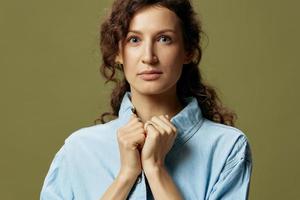 Serious adorable curly beautiful female in jeans casual shirt touches collar looks at camera posing isolated on olive green background. Being Yourself. People Lifestyle emotions concept. Copy space photo