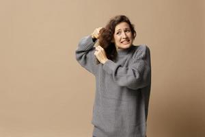 Tormented curly beautiful female in gray casual sweater with hairbrush comb pulls tangled hair posing isolated on over beige pastel background. Problematic unruly damaged hair concept. Copy space photo