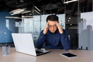 Stressed man working inside office, overworked asian man sitting at workplace sad and depressed holding hands on head, businessman frustrated thinking about problem photo