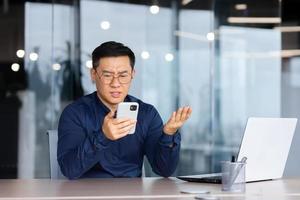 empresario leyendo malo Noticias en línea desde teléfono, asiático hombre decepcionado y triste mirando a teléfono inteligente pantalla, hombre en camisa trabajando dentro oficina utilizando ordenador portátil a trabajo foto