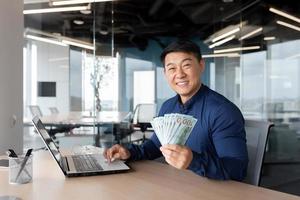 A young Asian man sits in the office with a laptop. He holds a phone and cash in his hands photo