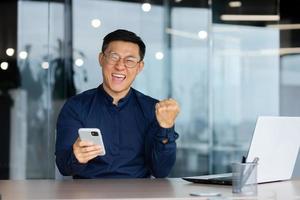 Portrait of happy Asian businessman, man celebrating achievement good results and victory, boss looking at camera inside office holding phone, entrepreneur working inside office photo