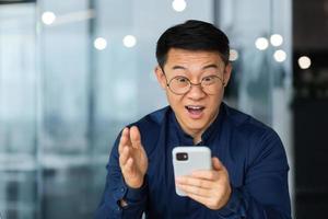 Close up photo of man in glasses and shirt shocked by good news smiling and looking at laptop screen, successful Asian businessman working inside modern office building