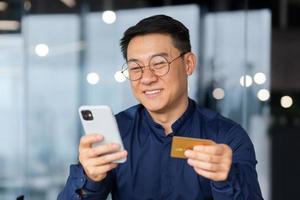 Successful and smiling asian businessman in office making bank transfer and online purchase in online store, man using app on phone and bank credit card, close up inside photo