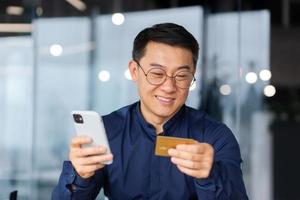 un contento joven asiático hombre es sentado en el oficina, participación un crédito tarjeta y teléfono en su manos foto