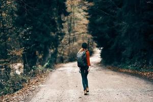 woman in jeans jacket with a backpack walks along the road in the autumn forest photo