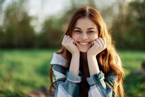 A beautiful woman outdoors in a green park laughs and smiles as she looks into the camera at sunset in the sun. Close-up portrait photo