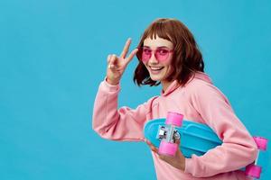Funny joyful pretty redhead lady in pink hoodie sunglasses with penny board smiling posing isolated on blue studio background. Copy space Banner Offer. Fashion Cinema. Holiday activity photo