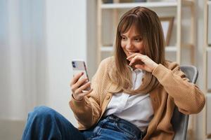 Cute young blonde lady in warm sweater doing selfie use phone hold hand near face sitting in armchair at modern home. Pause from work, take a break, social media in free time concept. Copy space photo