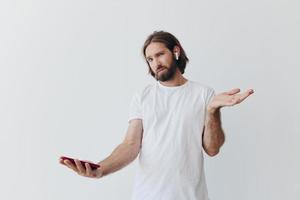A man with a beard blogger in a white T-shirt with a phone and wireless headphones looks into the phone and spreads his hands to the side on a white background isolated copy space photo