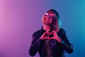 Lovely beautiful blonde woman in leather jacket sparkly dress trendy sunglasses show heart with fingers posing isolated in blue pink color light studio background. Neon party concept. Copy space photo