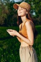 mujer blogger con teléfono en mano en naturaleza en contra un fondo de verdor sonriente en el Brillo Solar vistiendo un gorra después hacer ejercicio foto