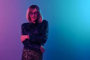 Excited sexy beautiful blonde woman in leather jacket sparkly dress trendy sunglasses cross hands posing isolated in blue pink color light studio background. Neon party Cyberpunk concept. Copy space photo