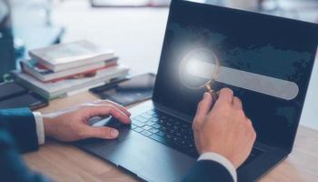 search engine concept, businessman working on computer laptop holding magnifying glass and search icon on busy table in office photo
