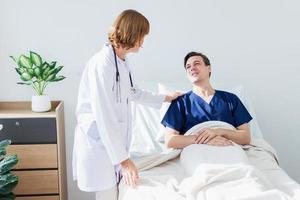 el médico es examinando el paciente en el hospital. caucásico hembra médico hablando a masculino paciente acostado en hospital cama. foto