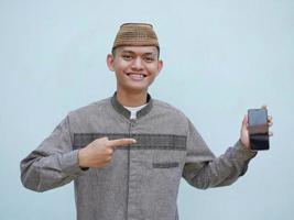 retrato de joven asiático musulmán hombre utilizando teléfono inteligente con contento expresión y señalando mano foto