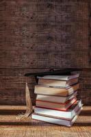 un académico gorra mentiras en parte superior de un apilar de libros en un de madera antecedentes. graduación y graduación concepto foto