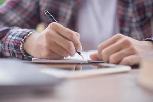 un negocio hombre es tomando notas dentro un cuaderno con un alarma reloj en el mesa. foto