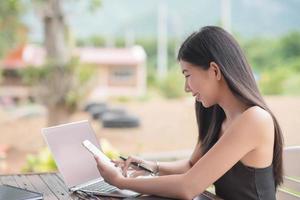 joven mujer de negocios difícil a trabajo a su escritorio escritura notas en un almohadilla desde su ordenador portátil computadora con analítico cartas y gráficos en frente de su, alto ángulo ver foto