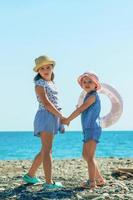 two little sister girls came to the sea in the summer, they are dressed in summer beach clothes, in panama hats and with an inflatable swimming ring photo