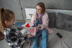 The daughter congratulates her mother on Mother's Day, gives her a box with a gift and a congratulatory opener with a heart made by herself. A woman knits in her bedroom. photo