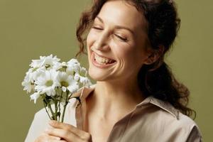 Happy cheerful curly beautiful female in linen casual shirt hold chamomiles enjoys flowers smell posing isolated on over olive green pastel background. Nature is beauty concept. Copy space photo