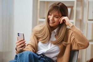 Cute young blonde lady in warm sweater doing video call with friends use phone sitting in armchair at modern home interior. Pause from work, take a break, social media in free time concept. Copy space photo