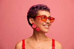 Moda retrato de un mujer con un corto Corte de pelo en de colores Gafas de sol con raro accesorios con pendientes sonriente en un rosado brillante antecedentes foto