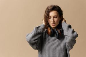 Serious calm adorable curly beautiful female in gray casual sweater headphones ready to enjoy fav songs posing isolated on over beige pastel background. Sound streaming platform concept. Copy space photo